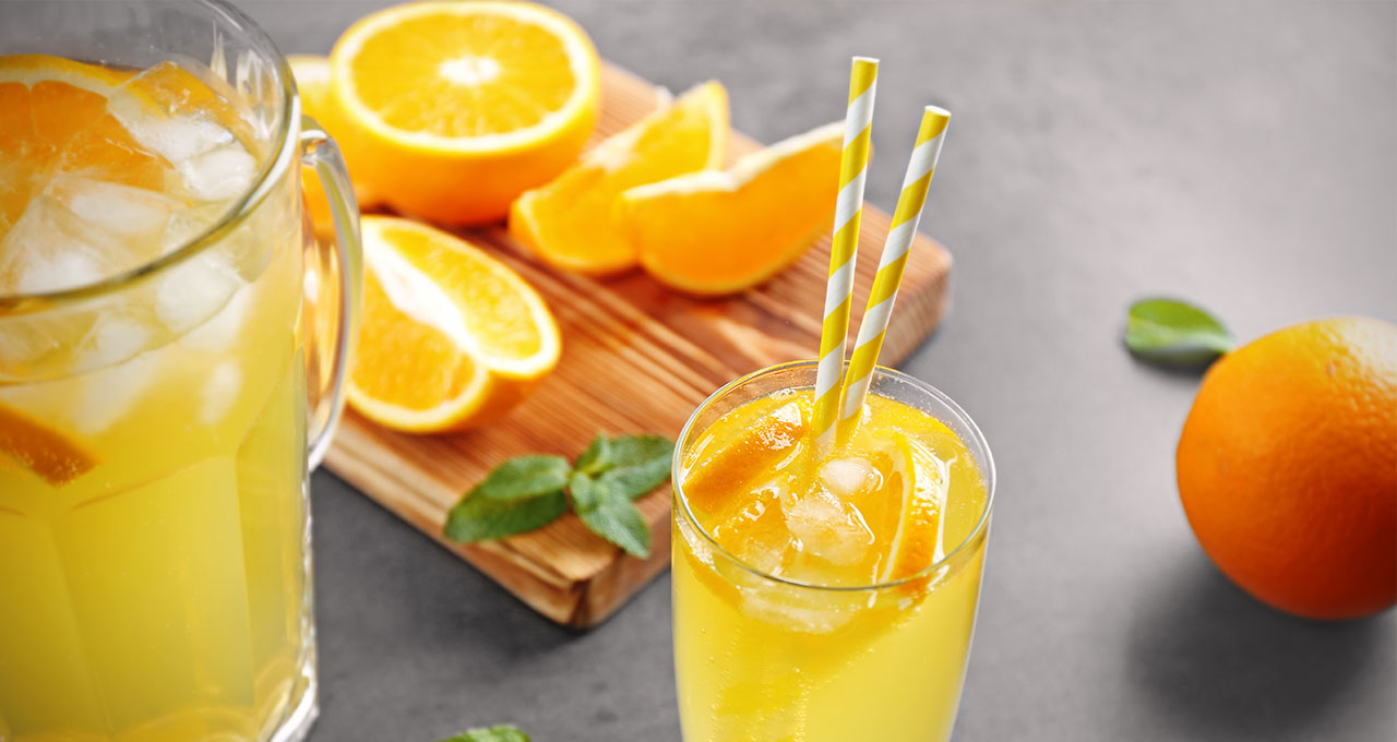 Orangenlimonade im Glas mit Eiswürfeln, Orangenscheiben im Hintergrund